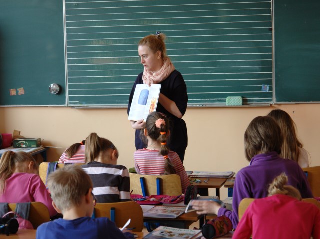 nauczycielka stoi obok tablicy, trzyma książkę w dłoni i coś tłumaczy dzieciom