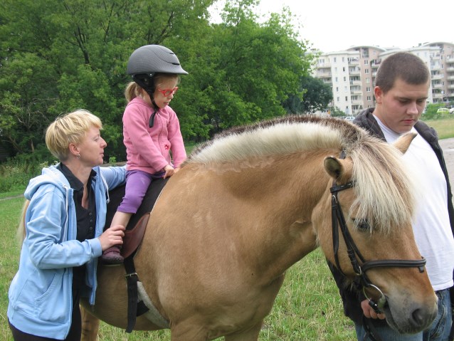 mała dziewczynka w różowej bluzie i oprawkach okularów jedzie na koniu przy trenerce i pomocnikiem 