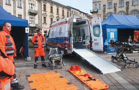 Karetka pogotowia, obok stoi dwóch ratowników, leżą nosze, karetka ma rozłożony podjazd