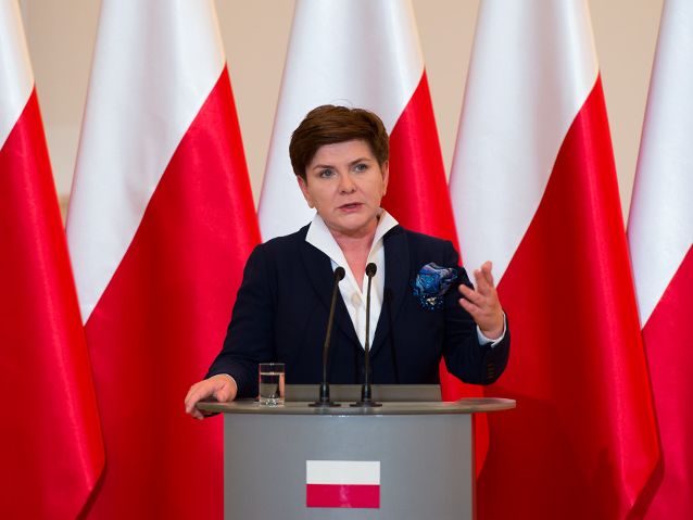 Premier Beata Szydło na mównicy, za nią flagi Polski