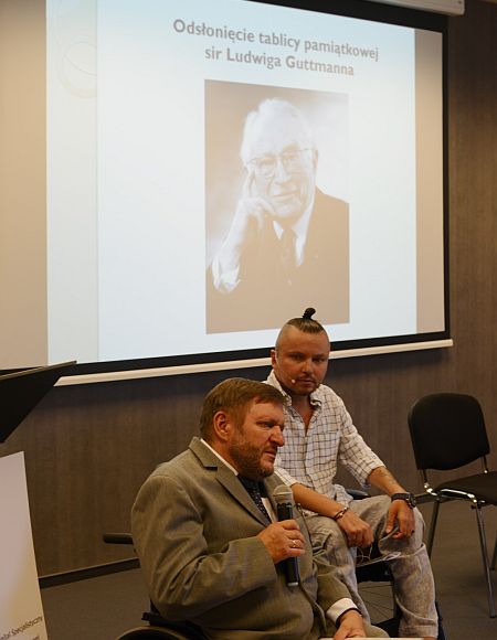 Konferencja, przy mikrofonie poseł Sławomir Piechota, za nim Bartłomiej Skrzyński, oba na wózkach, w tle zdjęcie Ludwiga Guttmanna
