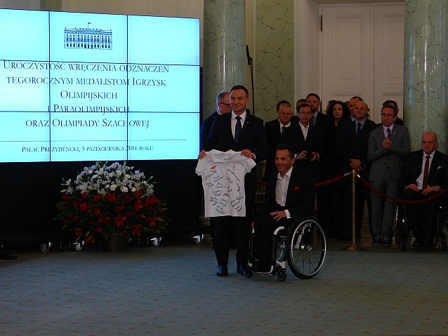 Reprezentacyjna sala w Pałacu Prezydenckim. Prezydent na środku stoi z koszulką w rękach, obok na wózku Rafał Wilk
