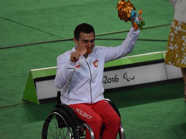 Adrian Castro uśmiechnięty, z brązowym medalem w jednej ręce i makotką Igrzysk w drugiej