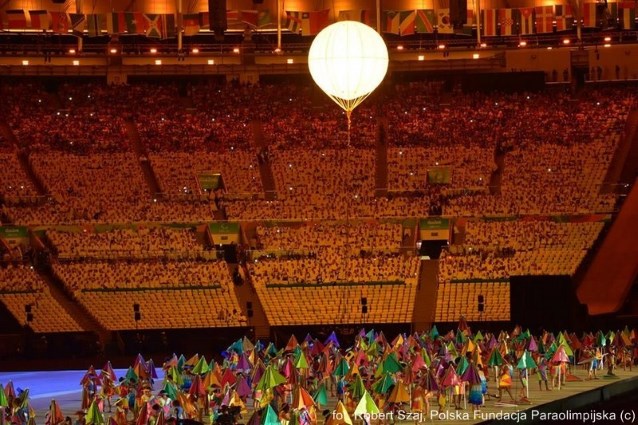 ponad kolorowym tłumem osób na scenie znajduje się balon, w tle kibice na stadionie