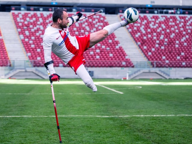 ampfutbolista podpierając się na kuli, w wyskoku odbija nogą piłkę