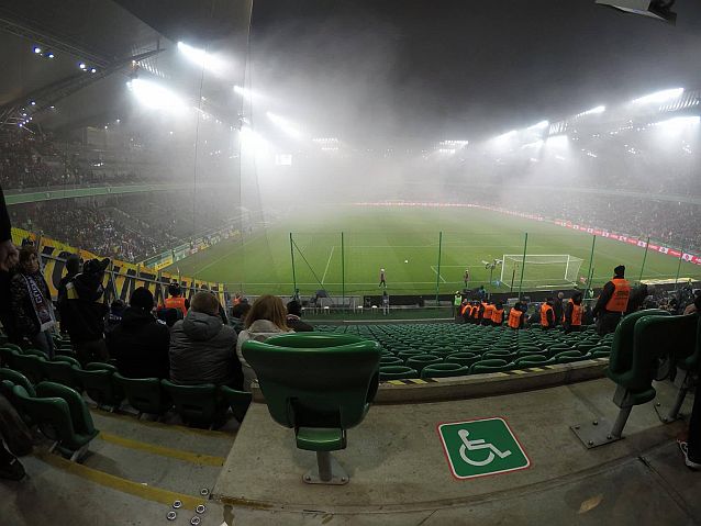 Stadion w czasie meczu, widok z trybun, na pierwszym planie miejsce oznaczone piktogramem wózka inwalidzkiego