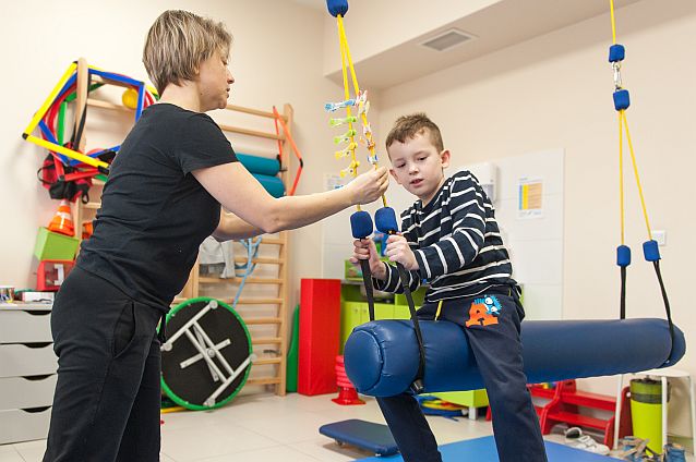 Sala ćwiczeń. Terapeutka ćwiczy z chłopcem, który siedzi na wiszącej miękkiej belce