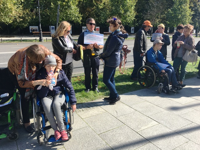 na pierwszym planie matka podaje wodę swojej córce na wózku, na drugim planie znajdują się protestujący rodzice