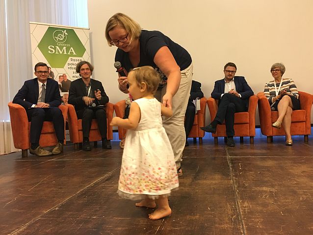 Mała dziewczynka idzie trzymana za rączkę przez mamę. W tle uczestnicy konferencji nt. SMA