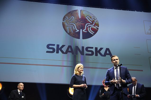 Adam Targowski przy mikrofonie stoi ze statuetką, obok niego stoi Joanna Ejsmont, w tle napis: Skanska