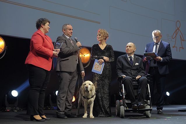 Paweł Wdówik z psem przewodnikiem na scenie przy mikrofonie, obok trzymająca statuetkę Donata Kończyk oraz prowadzący galę: Monika Zamachowska, Piotr Pawłowski i Michał Olszański