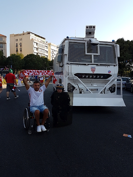 Łukasz Irla na wózku z uniesionym szalikiem reprezentacji Polski, obok policjant z tarczą i polewaczka do tłumienia demonstracji