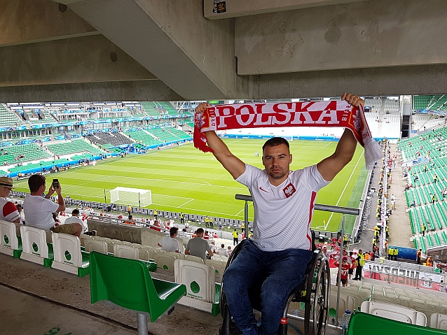 Siedzący na wózku Łukasz Irla z szalikiem uniesionym nad głowę dla tle trybun stadionu