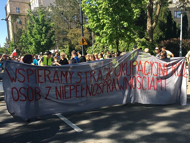 Czoło manifestacji. Parę osób niesie wielki transparent z napisem: wspieramy strajk okupacyjny rodziców osób z niepełnosprawnościami