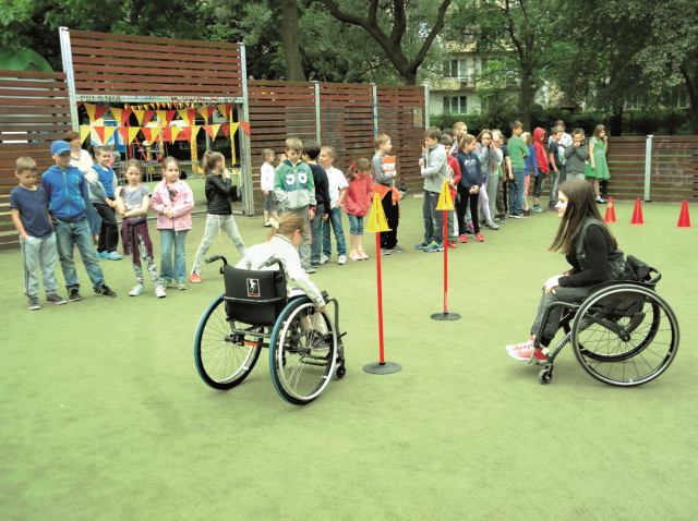 dziewczyna na wózku edukuje dziewczynkę, która usiadła na wózek jak się na nim poruszać. Obok stoją dzieci, czekając na swoją kolej