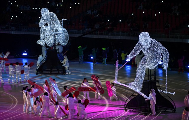 Ogromne figury, oświetlone lampkami, przedstawiają sportowca na wózku i sportowca bez nogi na narcie z kijkami. Poniżej figur stoją dzieci