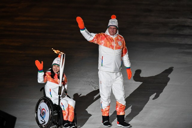 Koreanka na wózku ma przyczepiony do wózka drążek z ogniem paraolimpijskim, obok kobiety idzie jej trener