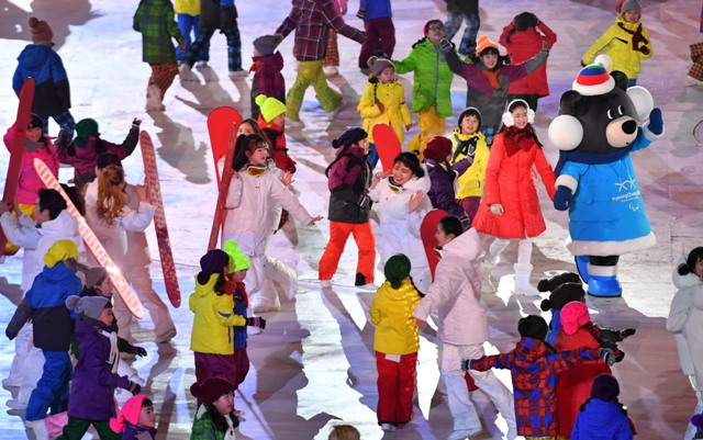 Dzieci w kolorowych kurtkach tańczą na płycie stadionu