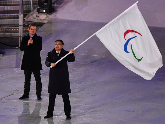 Chiński przedstawiciel trzyma w dłoniach flagę paraolimpijską, za nim klaszcze Andrew Parsons