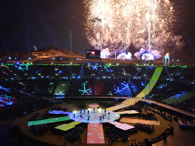 Na płycie stadionu tańczą maskotki Paraolimpiady, nad stadionem strzelają fajerwerki