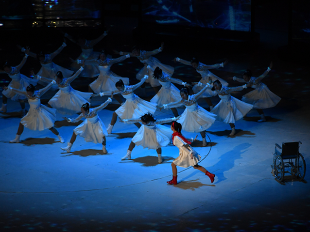 Kilkanaście koreańskich nastolatek w białych sukienkach tańczy, podbiega do nich dziewczyna w białek sukience, czerwonym szalikiem i w czerwonych butach. Obok nich stoi pusty wózek, z którego zerwał się tancerka 
