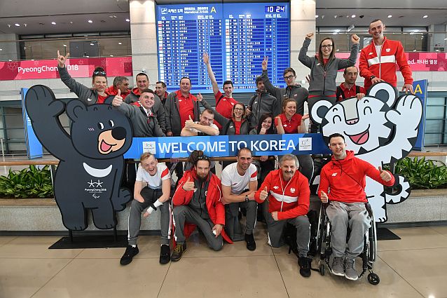 Reprezentanci Polski, trenerzy i działacze pozują uśmiechnięci do wspólnego zdjęcia na lotnisku w Pjongczangu. Napis na środku: welcome to Korea