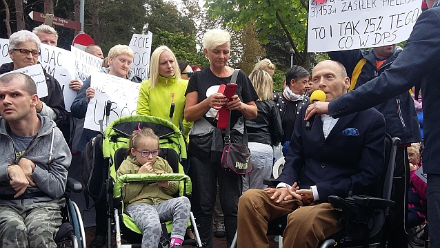 Manifestacja. Ludzie stoją z transparentami. Do mikrofonu mówi Piotr Pawłowski