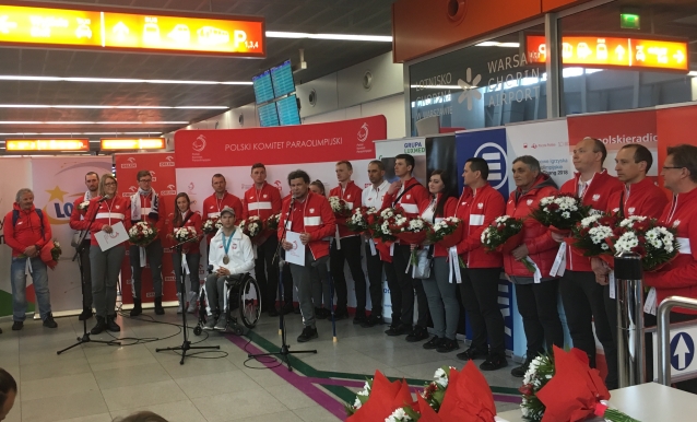 Paraolimpijczycy stoją z kwiatami na lotnisku. Przemawia prezes Polskiego Komitetu Paraolimpijskiego, Łukasz Szeliga
