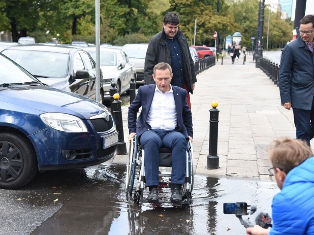 Paweł Rabiej przejeżdza przez dużą kałużę, tuż po zjechaniu z chodnika, by przejść przez ulicę. Za nim asekurant