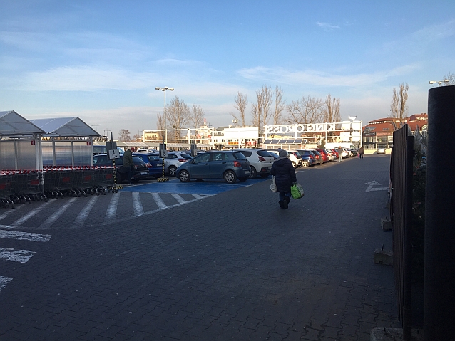 Parking zastawiony samochodami. Po lewej stronie wózki sklepowe pod wiatą, obok niej koperty dla osób z niepełnosprawnością