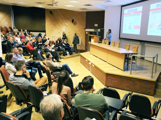 sala konferencyjna na której siedzą słuchacze przez mikrofon przemawia prelegentka