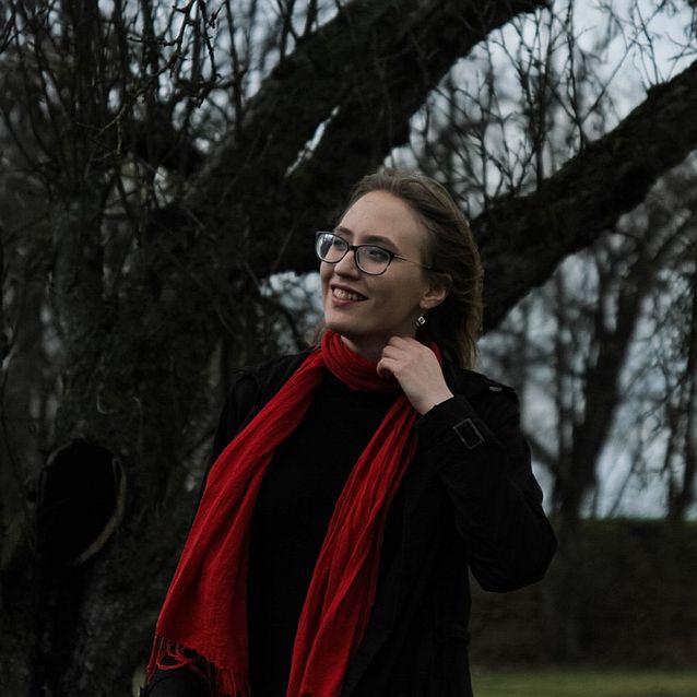 Weronika Ławicka w czarnym płaszczu i czerwonym szaliku pozuje do zdjęcia na tle drzew