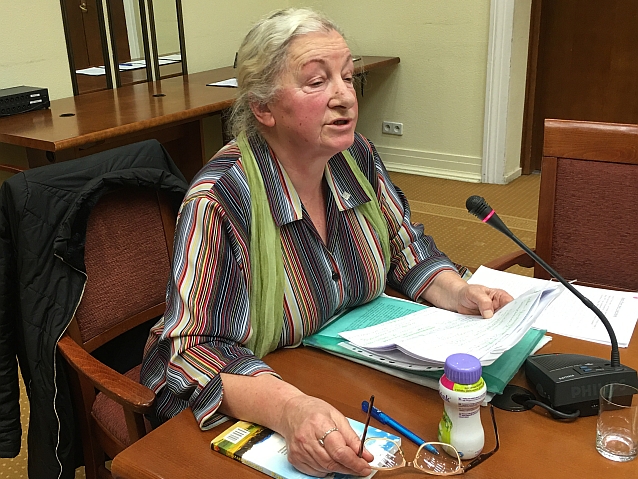 Starsza kobieta siedzi za stołem, trzymając notatki i mówi do mikrofonu