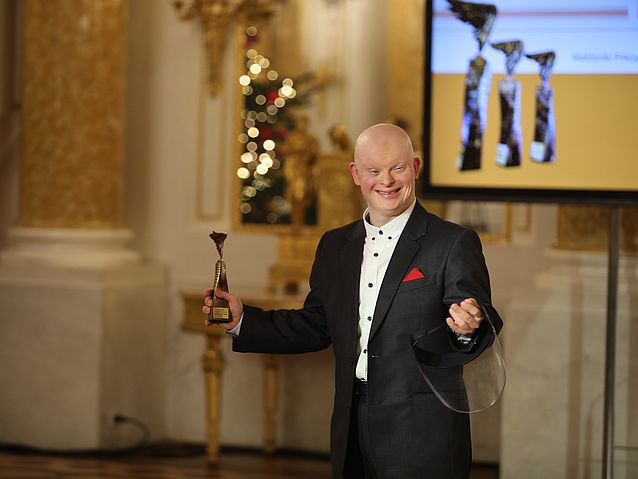 Uśmiechnięty, elegancko ubrany Michał Milka stoi trzymając statuetkę