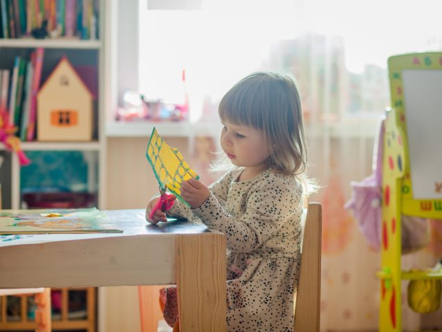 kilkuletnia dziewczynka siedzi sama przy stoliku i wycina nożyczkami z kolorowego papieru