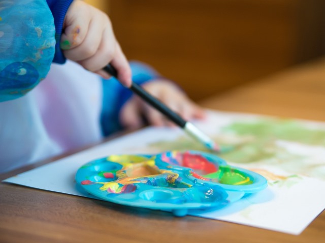 dziecko trzyma pędzelek w ręku nad kartką papieru leżą farbki