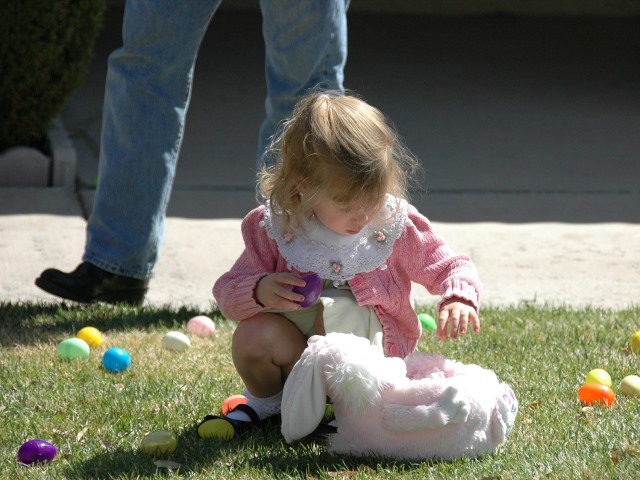 dziewczynka w kucki na trawie przy leżącej maskotce wokół plastikowe jajka za nią nogi taty