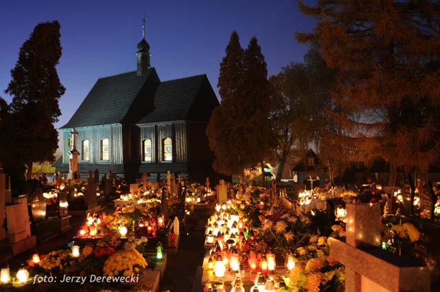 cmentarz oświetlony zniczami, w tle drewniany kościółek