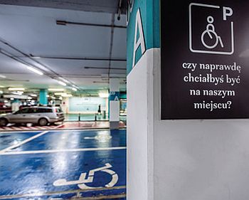 Parking podziemny. Niebieskie tzw. koperty. Na filarze symbol osoby na wózku i napis: czy naprawdę chciałbyś być na naszym miejscu?