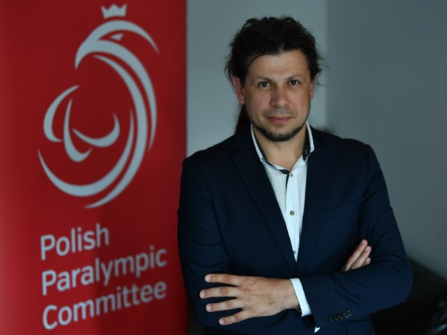 Łukasz Szeliga stoi z założonymi rękami na tle czerwonego rollapa z napisem polish paralympic committee