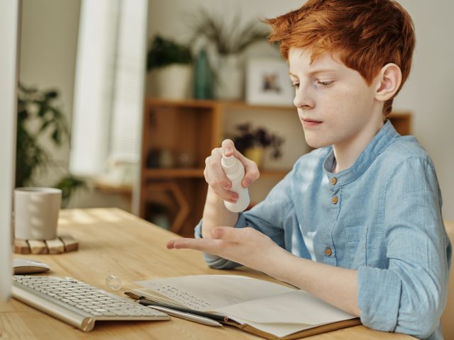 Chłopiec ok. 12-letni z rudymi włosami siedzi przy stole leży zeszyt i klawiatura a on pryska sobie ręce płynem dezynfekującym