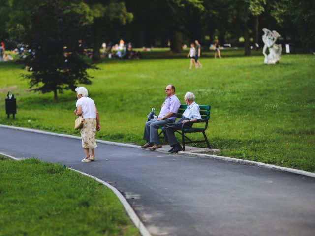 w parku starsza kobieta idzie ścieżką na ławce siedzi dwóch seniorów