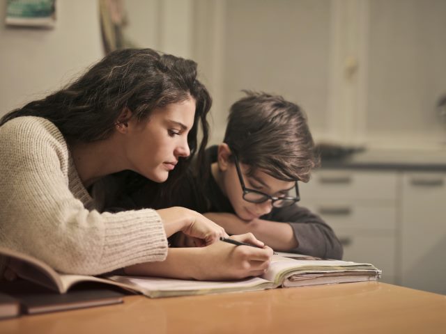 kobieta pochylona nad zeszytami obok z długopisem w dłoni chłopie w okularach, siedzą przy stole