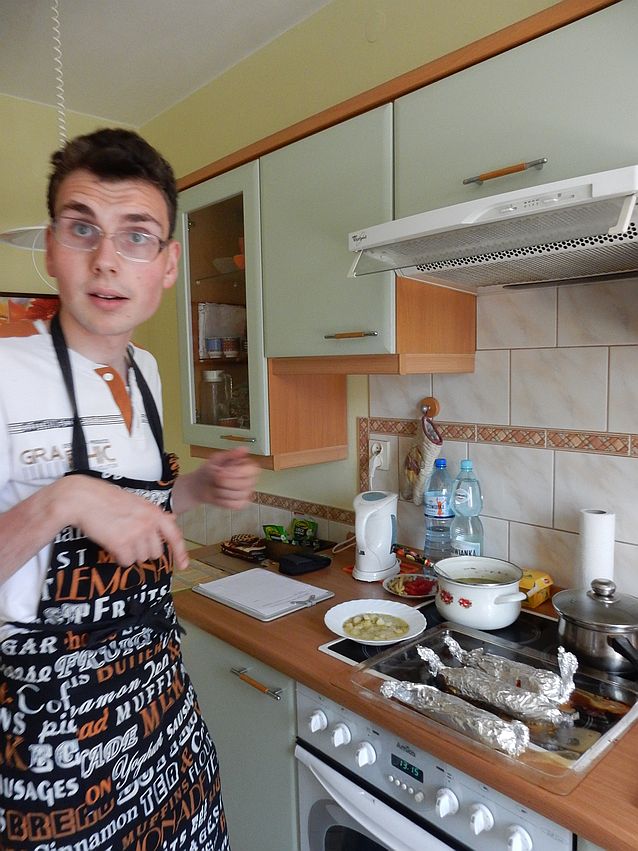 Tomasz Sikora w fartuchu kuchennym stoi w kuchni przy kuchence, na której stoją garnki i trzy przedmioty zawinięte w folię