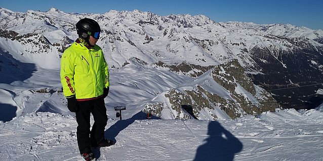 Wojciech w kurtce narciarskiej, kasku i goglach, ale bez wpiętych nart, na tle zaśnieżonych gór