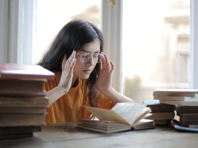 dziewczyna z długimi włosami w okularach siedzi przy stole zawalonym książkami ma zamknięte oczy i obie ręce opiera w okoliach uszu