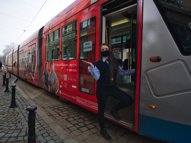 motornicza w maseczce z jedną nogą na stopniach wejścia do tramwaju i wyciągniętą jedną ręką w geście zaproszenia