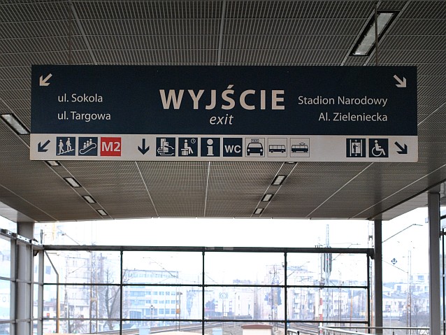 Wisząca na górze tablica informacyjna na stacji kolejowej z dużym napisem wyjście, strzałkami do poszczególnych ulic i ikonkami