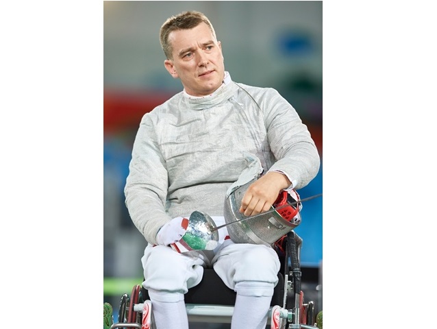Grzegorz Pluta na wózku w stroju sportowym z szablą i przyłbicą