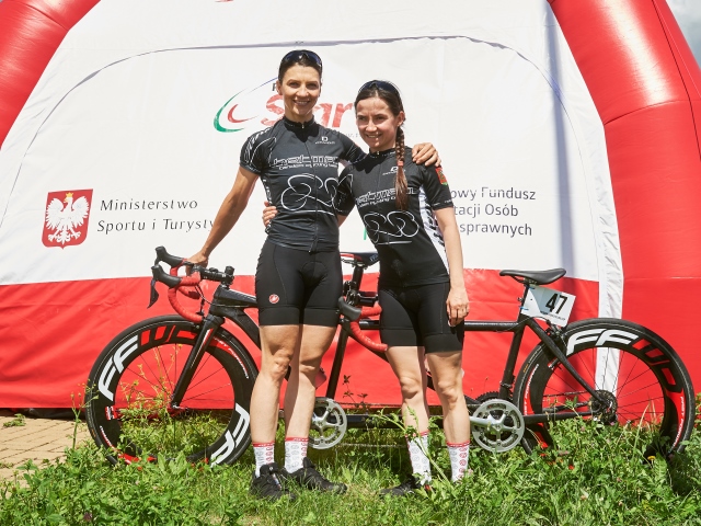 Kiryła i Tecław objęte uśmiechają się do obiektywu w tle rower i namiot z logo startu